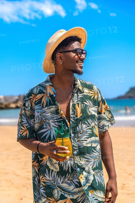 Young man wearing a colorful hawaiian shirt and stylish sunglasses, enjoying a refreshing pineapple shaped cocktail on a sunny beach