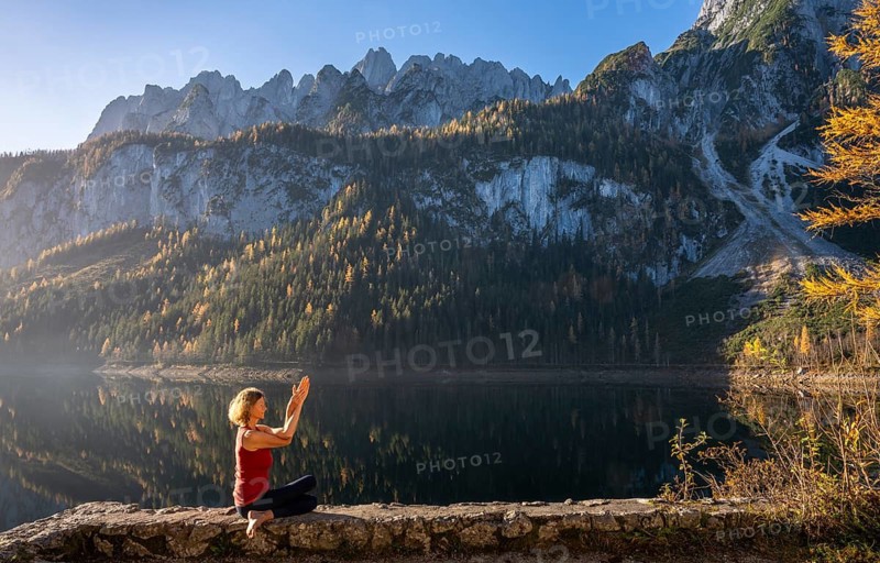 A woman doing yoga (Gomukhasana, Garudasana) by the lake. Vorderer Gosausee in autumn with a view of the Dachstein mountains. The Gosaukamm on the right. Blue sky, good weather. Reflection. Morning atmosphere. Vorderer Gosausee, Gosau, Gosau valley, Salzkammergut, Upper Austria, Austria