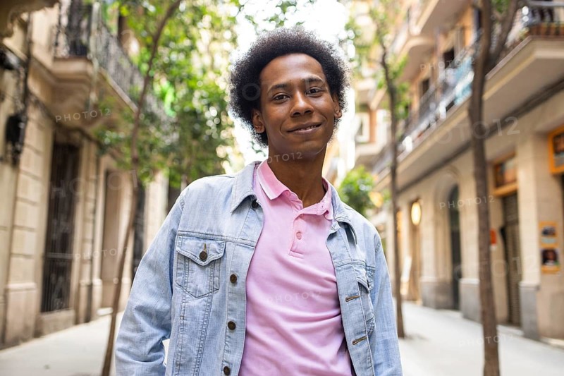A non-binary person of African American descent walks confidently down a sunlit urban street, wearing a light denim jacket. The scene captures diversity and individuality