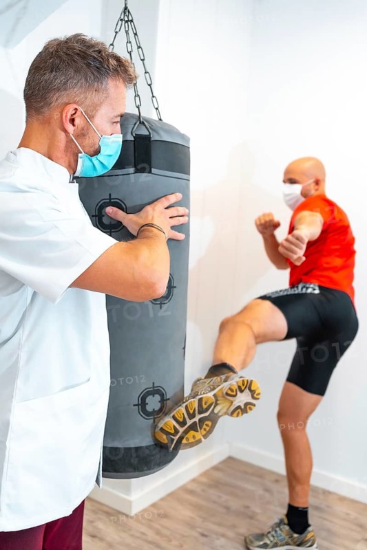 Physiotherapist with mask together with the patient on the punching bag. Physiotherapy with protective measures for the Coronavirus pandemic, COVID-19. Osteopathy, sports quiromassage