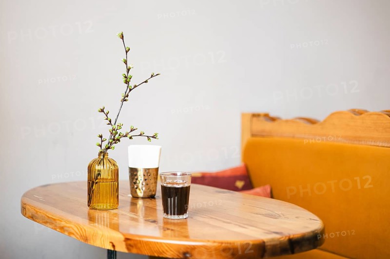A cozy cafe scene featuring a wooden table with a glass of coffee, a textured cup, and a simple vase with delicate branches. The setting is warm and inviting