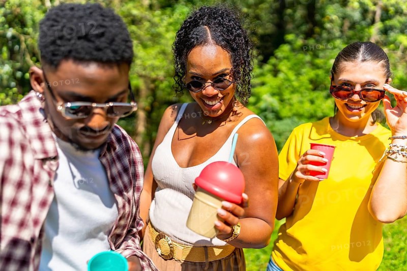 Three friends are having refreshing drinks together on a sunny day in a park, enjoying each other's company and the pleasant weather