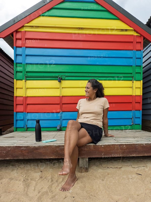 Colorful Brighton Bathing Boxes in Melbourne, Australia, with a person relaxing in front of a vibrant, rainbow-painted beach hut, enjoying the sunny day at the seaside