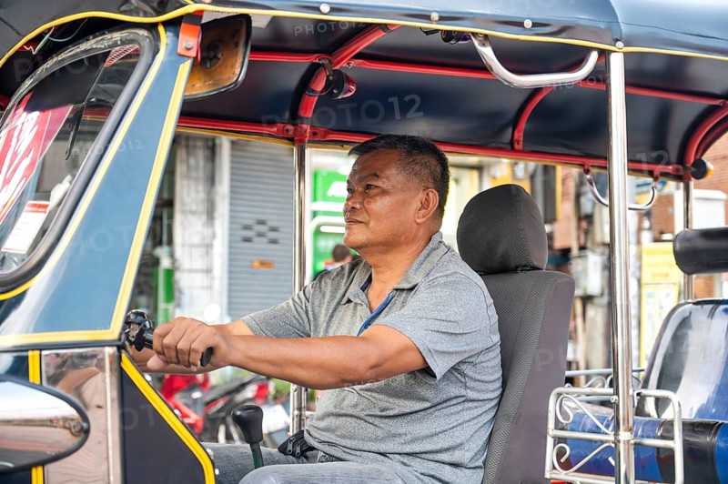 A Thai male tuk tuk driver sits in his vehicle, attentively awaiting passengers on a vibrant street in Chiang Rai. He appears focused and prepared for the day ahead