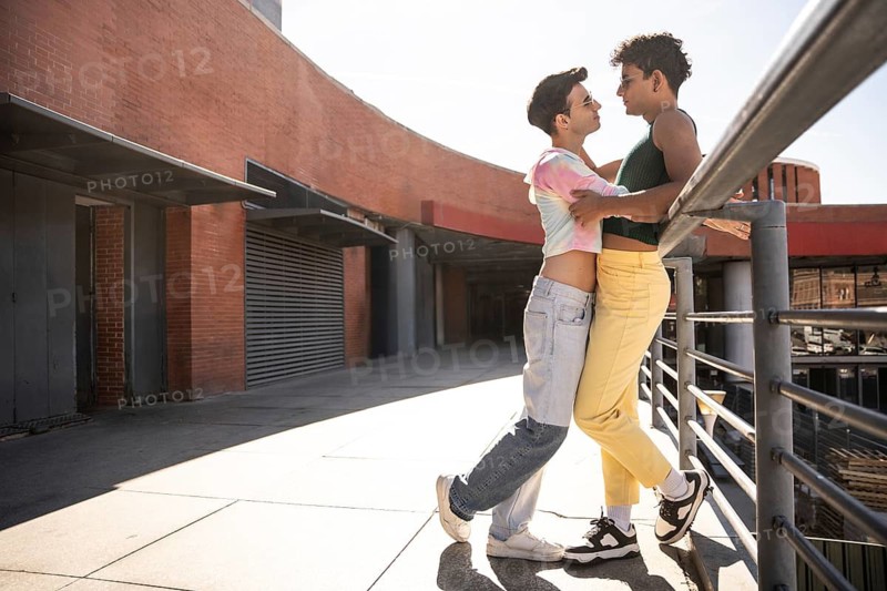 A gay couple shares an intimate embrace on a sunlit urban sidewalk near a red brick building. The scene captures a moment of connection in a modern cityscape