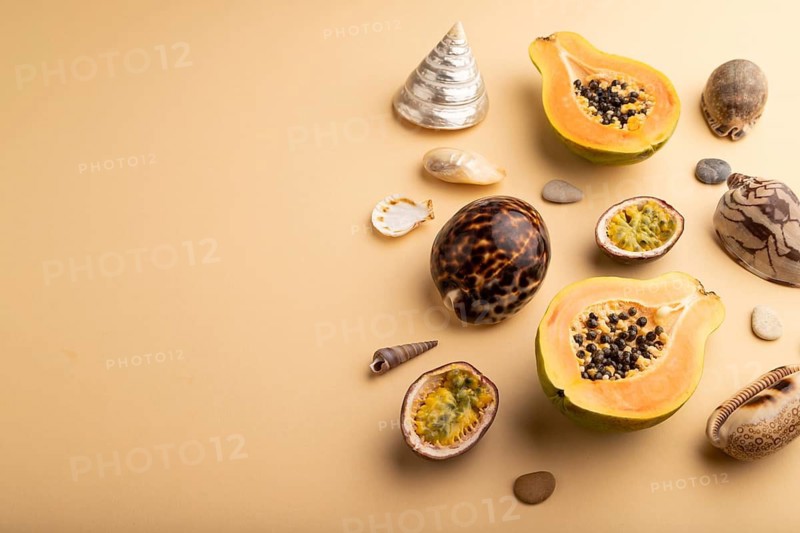 Ripe cut papaya, passion fruit, seashells, pebbles on orange pastel background. Side view, copy space. Tropical, healthy food, vacation, holidays concept
