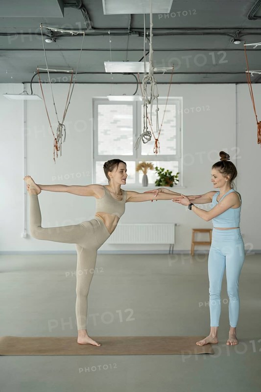 Yoga instructor guides a woman in performing a balance pose during a class. The serene studio setting enhances the focus on alignment, posture, and form