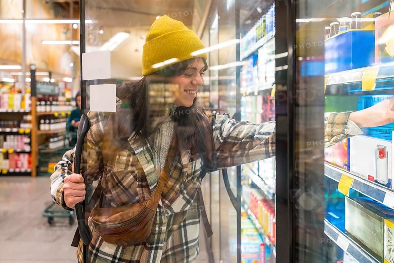 Young woman wearing a beanie and plaid shirt smiling while opening a refrigerated door to select an item from a grocery store cooler, showcasing everyday city life activities