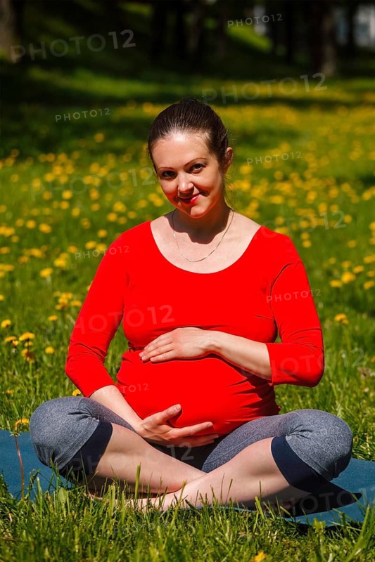 Pregnancy yoga exercise, pregnant woman doing asana Sukhasana easy yoga pose holding her abdomen outdoors on grass lawn with dandelions in summer