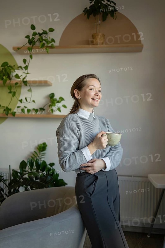 A woman stands in a stylish cafe, smiling and holding a cup of coffee. The bright, modern interior features plants and wooden shelves, creating a relaxing atmosphere