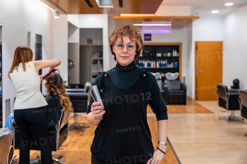 Professional hairdresser standing confidently in her salon holding a comb and clipper while a colleague styles a client in the background, showcasing modern beauty business life