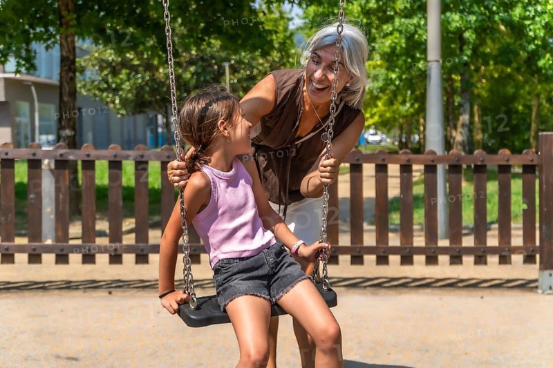 Granddaughter enjoying a fun swing ride at a park playground as her happy grandmother pushes the swing, sharing a joyful multigenerational family bonding moment outdoors during summer