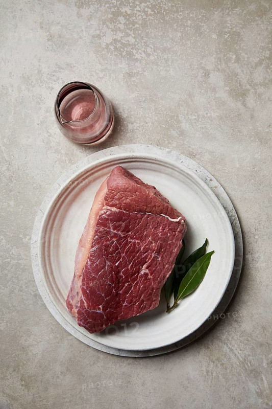 A fresh, raw beef steak placed on a ceramic plate, accompanied by aromatic bay leaves and a glass of rose wine, exemplifying a classic butcher theme