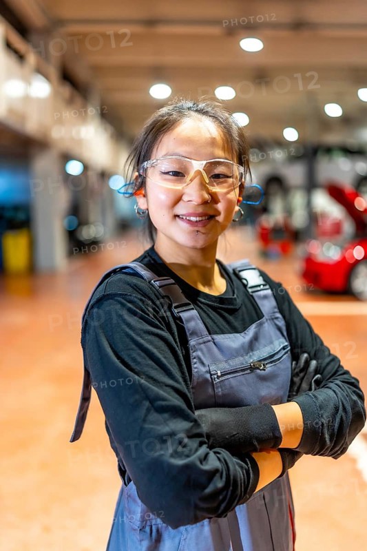 Confident female mechanic wearing protective glasses and earplugs, smiling with arms crossed in a car workshop