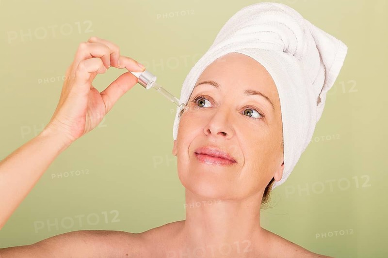A woman with a towel wrapped around her head applies serum to her face, exemplifying a self-care and skin care routine. The calm expression and soft background create a serene atmosphere