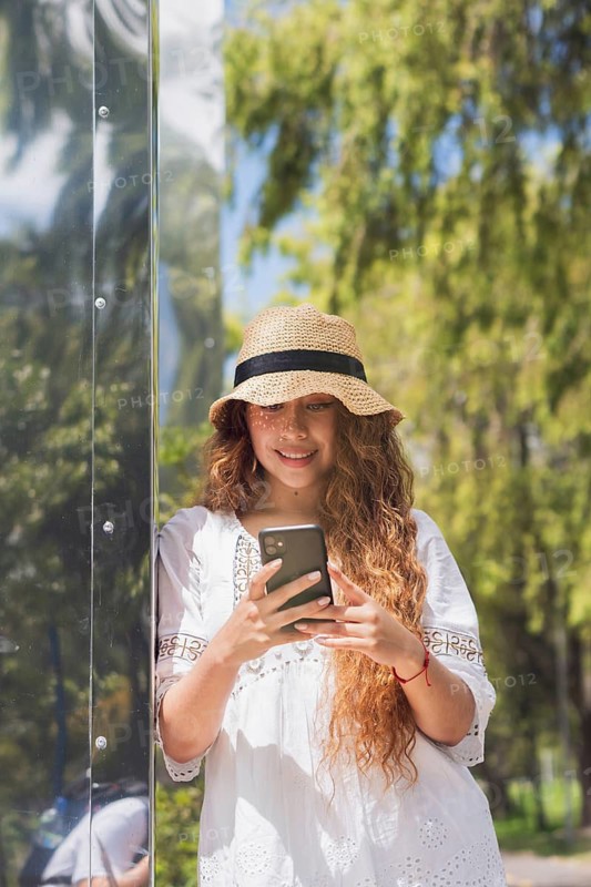 A young American Latin woman enjoying a sunny day outdoors, wearing a casual outfit and straw hat, while using her smartphone, standing against a reflective surface