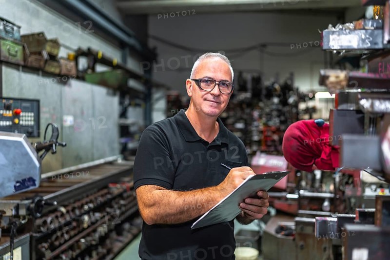 Mature male worker doing inventory in a metal cnc factory holding clipboard while looking at camera seriously