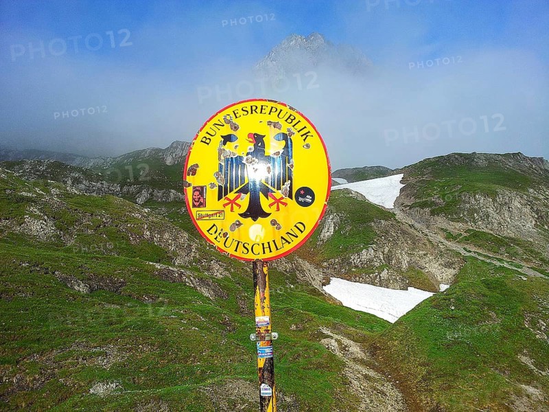 Yellow border sign of the Federal Republic of Germany against foggy mountain landscape with remnants of snow and green hills, mountain range in the background, E5, hiking in the Alps, crossing the Alps, Germany