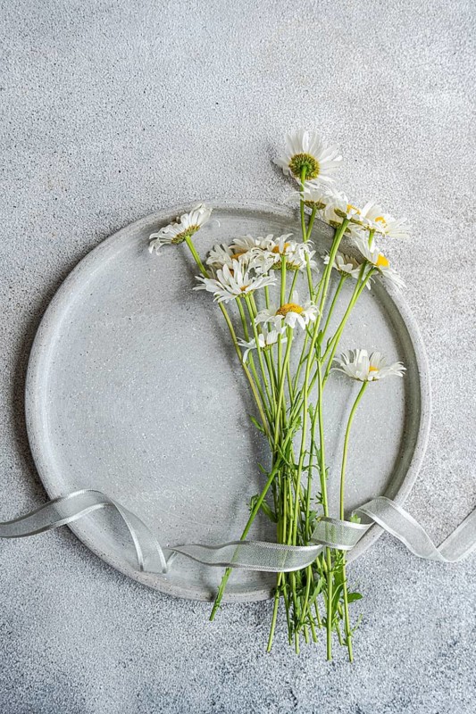 Top view of delicate daisy flowers arranged on a round ceramic plate, set against a textured concrete background. A subtle ribbon adds elegance to the composition