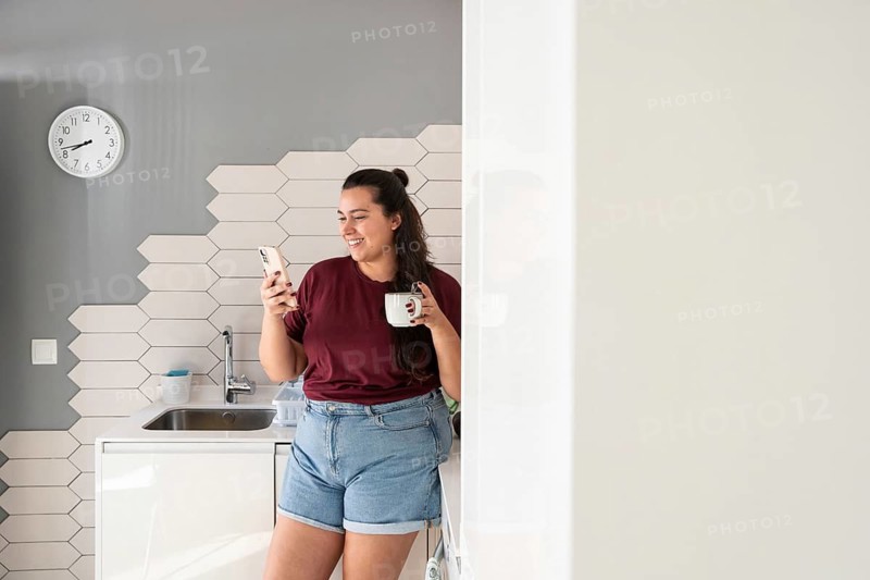 A curvy woman stands in a modern kitchen, smiling as she looks at her smartphone. She leans casually against the counter, enjoying a relaxed moment