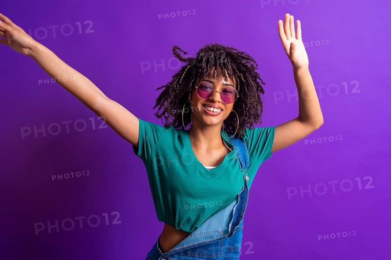 Carefree young woman with curly hair and sunglasses, wearing a green t shirt and denim overalls, dancing joyfully against a vibrant purple backdrop