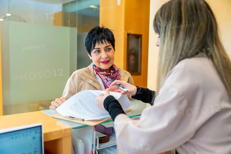 Mature woman smiling while completing paperwork requested by a female receptionist at the counter of a modern aesthetic or medical clinic, preparing for a consultation