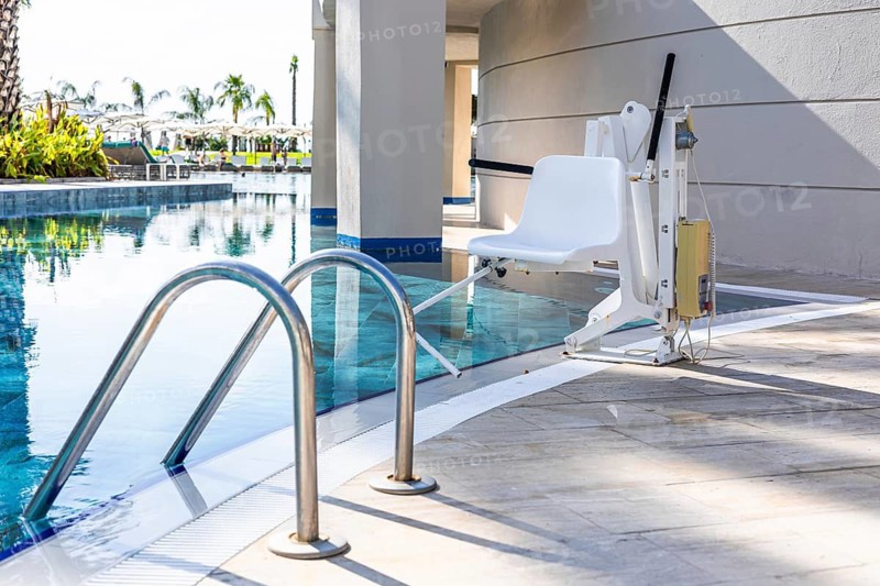 A poolside lift chair for disability people is positioned next to a swimming pool. The area is well-lit with clear blue water and a modern architectural design