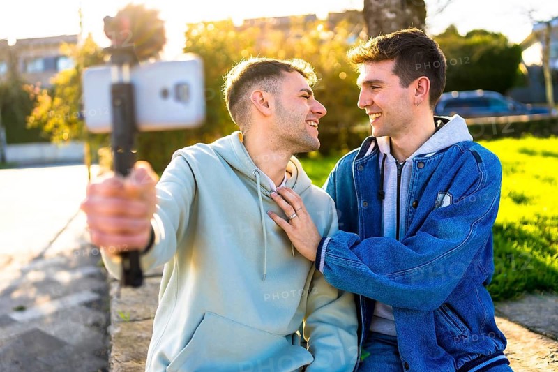 Smiling gay couple taking selfie outdoors using smartphone and gimbal, enjoying sunny day in urban park