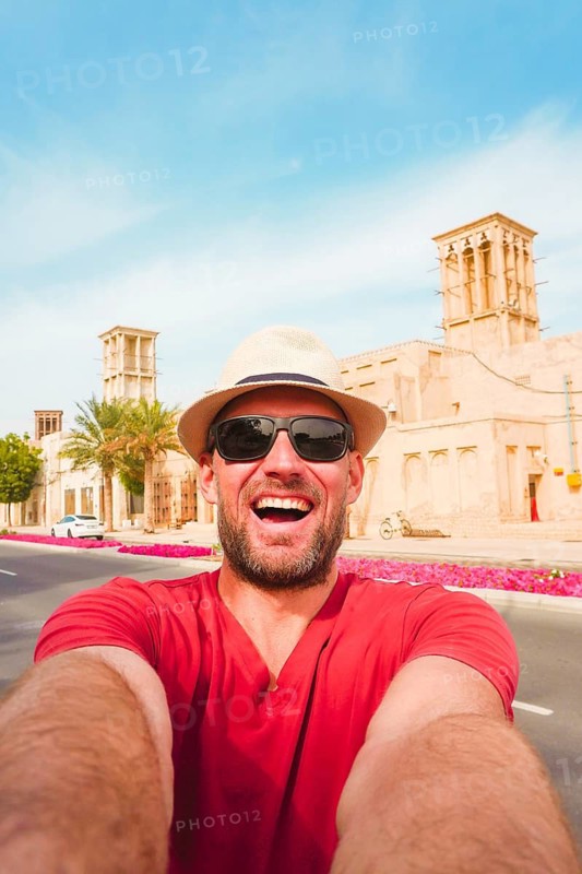 Happy male tourist wear red shirt and straw hat pose takes selfie at the Al Fahidi Historical Neighborhood in Old Dubai. Features a traditional wind tower backdrop cityscape view