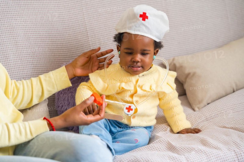 Horizontal cropped photo of an African little child playing doctors with her mother on the sofa