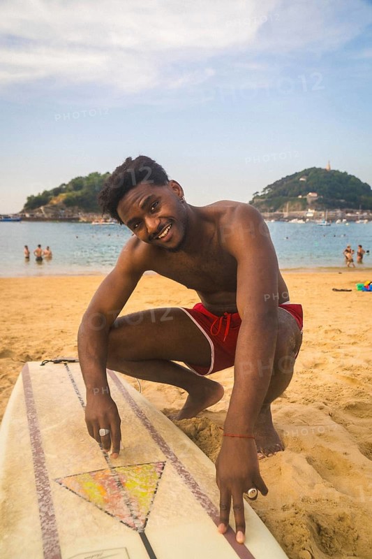 Young black man smiling at camera, crouching next to a surfboard on the sandy beach of la concha, san sebastian, enjoying a summer day and beach surfing lifestyle with an analog film aesthetic