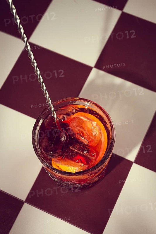 A drink fills a glass on a checkered table. Ice cubes and orange slices add color to the drink. A stirring tool rests in the glass