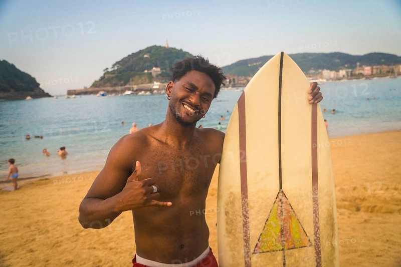 Young black man smiling and showing a shaka hand sign while standing with his surfboard on the sandy beach, enjoying summer vibes in san sebastian with people swimming in the ocean