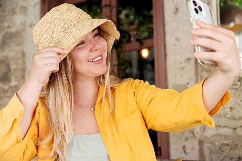 A young, cheerful woman wearing a yellow shirt and hat takes a selfie with her smartphone while sitting alone at a cafe table, capturing a moment of joy and leisure