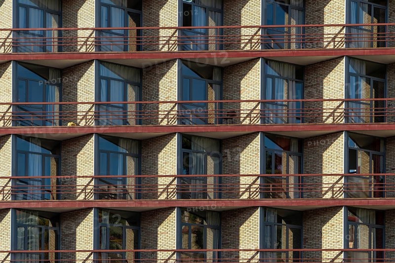 The image captures a modern apartment building with symmetrical balconies, showcased under bright sunlight, featuring red railings and beige brickwork
