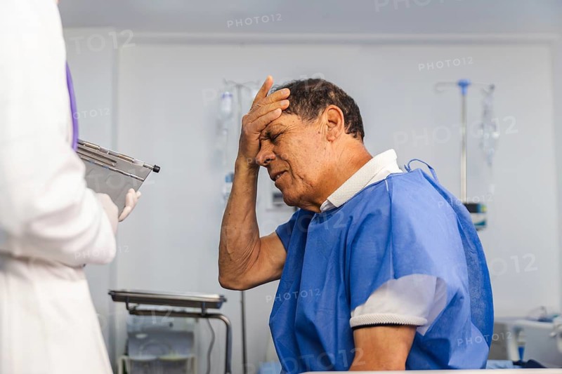 An elderly patient in a blue gown sits in a hospital room, holding his head in pain while consulting with a female doctor, who holds a clipboard during the medical assessment