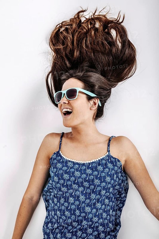 Woman wearing a blue patterned dress and sunglasses, lying on a white background. Her hair is artfully spread out, displaying a lively and joyful expression