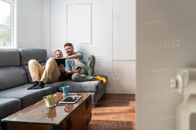 A cheerful gay couple relaxing on a modern gray sofa, engaging with a book. They enjoy quality time together in a cozy, bright living room setting