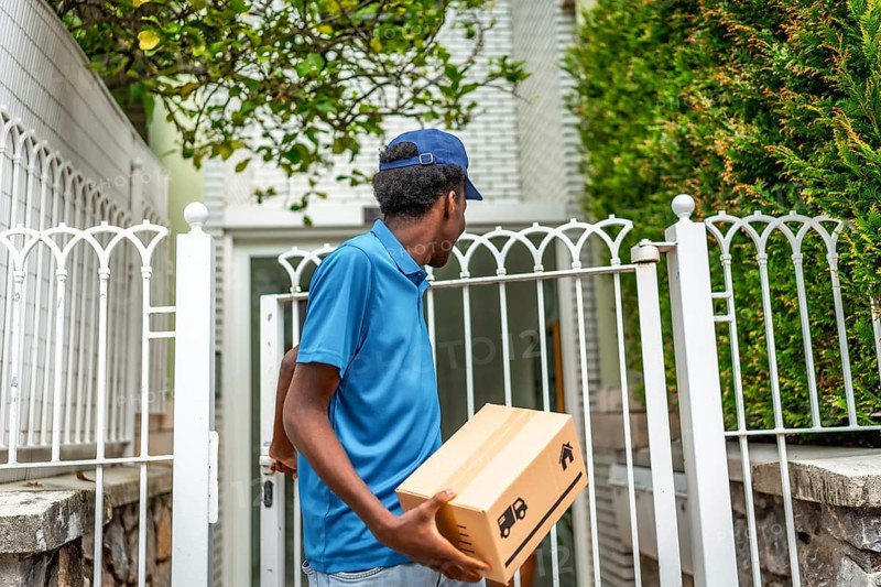 Rear view of an african courier delivering a package standing next to the entrance of a residential house