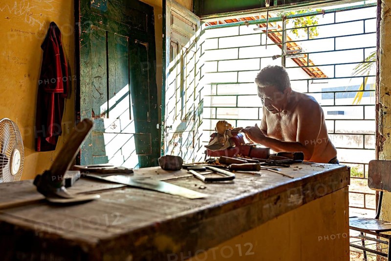 Man carving at a workbench in daylight of an open space using various tools, craftsman leg carving masks in Petrolina Brazil