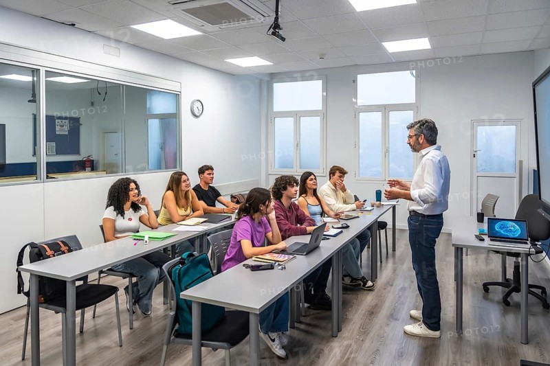 Teacher at front of bright classroom explaining lesson to diverse young adult students seated with laptops, engaging in discussion and attentive modern university learning