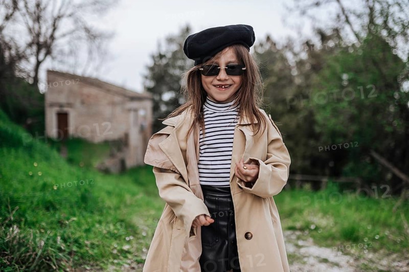 A young girl in Civitanova, Italy, wears a stylish outfit with sunglasses and a beret. She stands on a path surrounded by lush greenery and rustic buildings, exuding charm