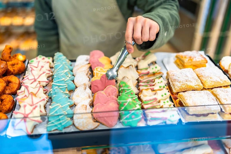 Close-up of a male baker using tongs to place an assortment of colorful Christmas pastries on a tray at the shop counter