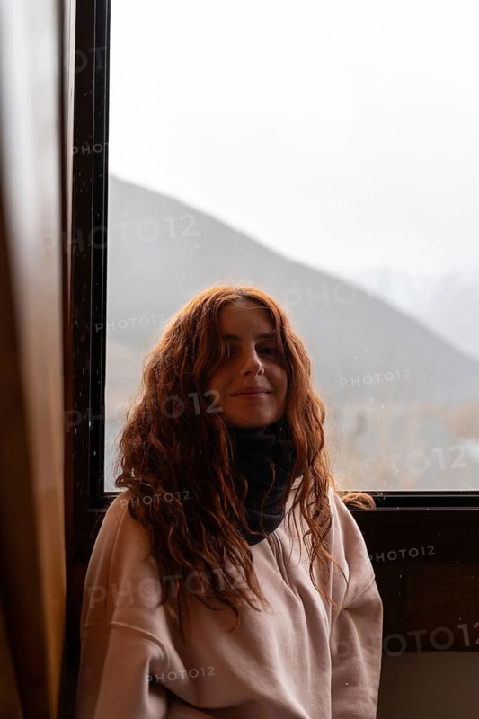 A young woman with red hair and a gentle smile is pictured comfortably inside during winter. She wears a soft sweater and scarf, gazing softly towards the camera