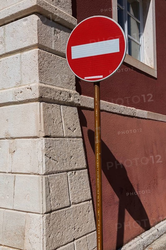 Red and white circular Do Not Enter pictogram notice sign for motor vehicle circulation attached to an old rusted steel post on sidewalk next to an unusual architectural style building at a street corner, Zadar, Croatia