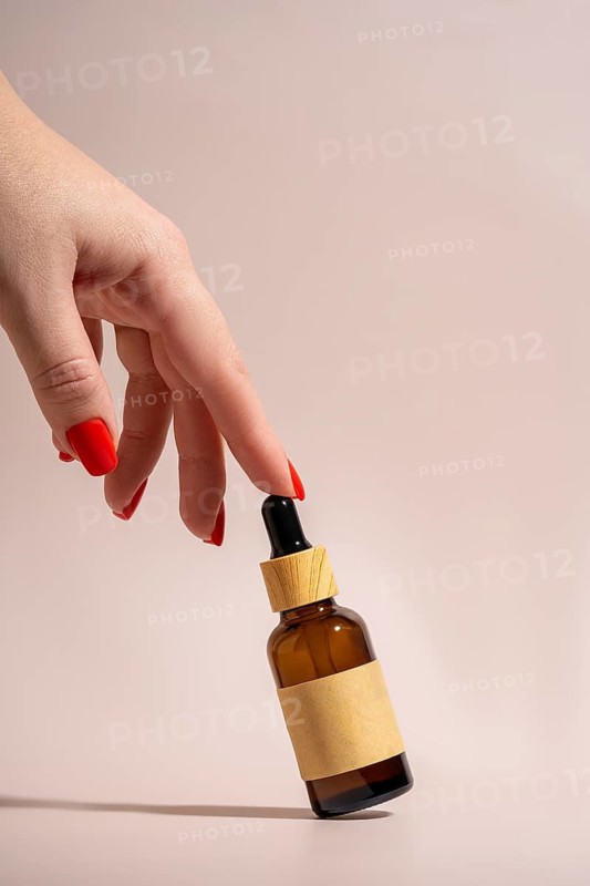 A hand with vibrant red nails elegantly holds an cosmetic amber glass dropper bottle against a soft beige background, evoking themes of beauty, skincare, and minimalism