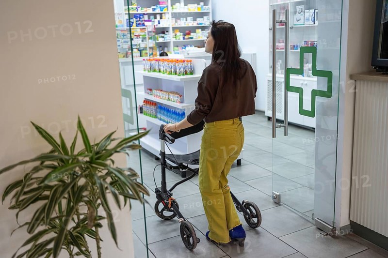 A woman with cerebral palsy using a walker enters a pharmacy, highlighting accessibility and independence in healthcare. The bright interior and green cross symbolize medical facilities