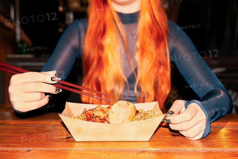 A woman with long red hair holds chopsticks over a tray of Asian street food, capturing the vibrant and diverse culture of an Asian food market