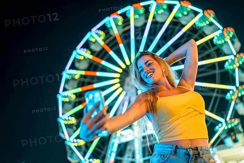 Low angle view of a beauty woman taking night selfie in an amusement park with an illuminated ferris wheel on the background