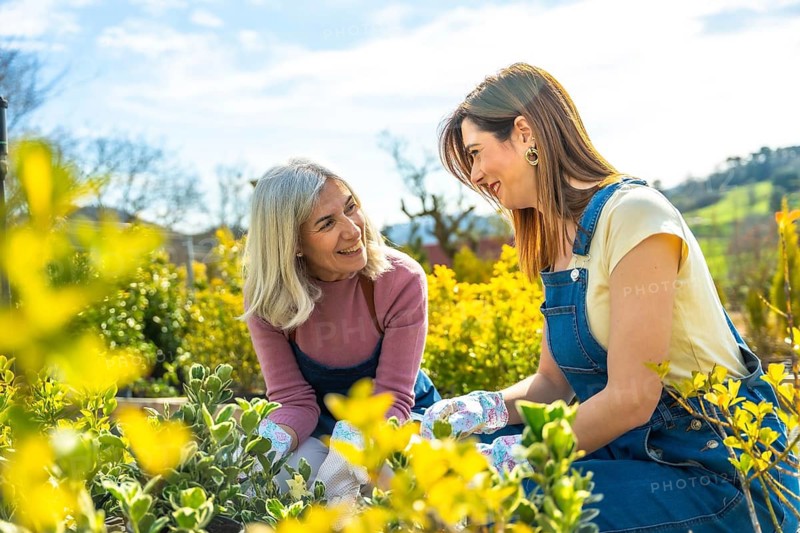 Grandmother and granddaughter smiling together while planting young green seedlings in pots, sharing gardening tips and enjoying sunny outdoor bonding and learning moments